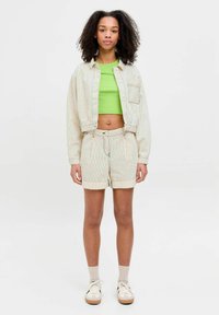 Giacca a righe color crema con tasca a toppa, abbinata a pantaloni corti coordinati. Un crop top verde brillante, sneakers chiare e calzini beige completano il look.