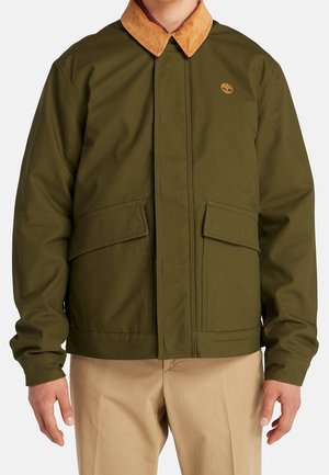 Olijfgroene jas met bruine ribfluwelen kraag, voorzakken met kleppen en Timberland-logo, gedragen met beige broek.