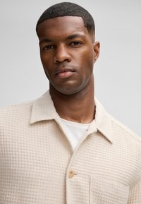 Camicia di colore crema con una texture a nido d'ape morbida, colletto e chiusura a bottoni, indossata sopra una maglietta bianca.
