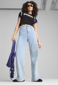 Camiseta negra corta con logo blanco de "PUMA", pantalones vaqueros de tiro alto y pata ancha en azul, y una mochila morada de cuerda. Modelo usando gafas de sol.