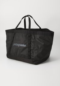 Fekete nylon bevásárlótáska két fogantyúval, sima felületű és gyűrt díszítéssel. A "Patagonia" logó szürkében az elején.
