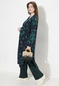 En lång, mörkblå cardigan med gröna blommönster, som är kombinerad med mörkgröna vida byxor och en liten vävd väska med ett handtag av trä.