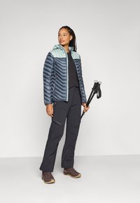 Jachetă căptușită în nuanțe de bleumarin și verde mentă, cu fermoar pe față, asortată cu pantaloni negri. Ținând bețe de trekking, purtând bocanci de drumeție maro.