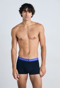 Boxers noirs en coton avec une ceinture élastique bleue comportant le texte "TOMMY HILFIGER". Le matériau semble lisse avec un design ajusté.