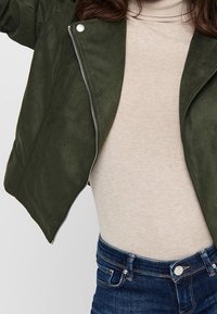 Veste cropped en faux suède olive avec détails de zipper en argent, superposée sur un col roulé beige, associée à un jean en denim bleu.