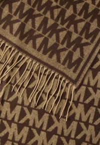 Barna mintás takaró, amely ismétlődő logó mintát tartalmaz kontrasztos árnyalatban, bézs zsinórokkal díszítve az egyik szélén.