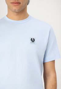 Τιρκουάζ κοντομάνικη μπλούζα από βαμβάκι, με τετράγωνο μαύρο και λευκό λογότυπο στο αριστερό μέρος του στήθους. Λείο υφή.