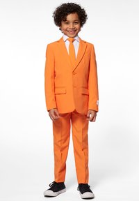 Orange jakkesæt lavet af struktureret stof, med en enkelt knap, to frontlommer, hvid skjorte og matchende slips. Sorte sneakers fuldender outfittet.