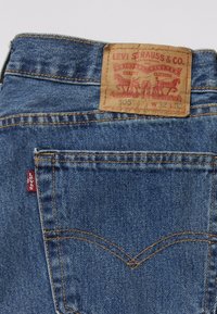 Levi's 505 traperice s tan kožnim označavanjem, plavim traperom, kontrastnim žutim šavovima i malom crvenom oznakom na stražnjem džepu.