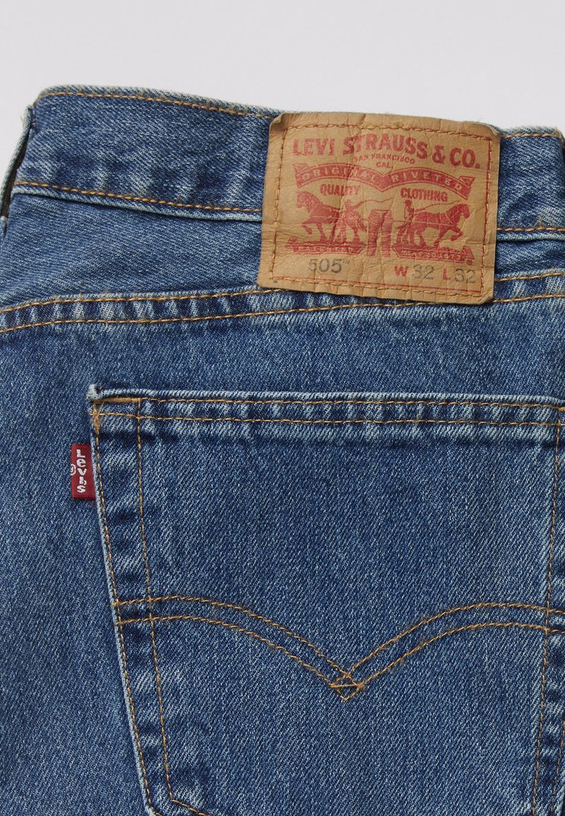 Jeans Levi's 505 avec un patch en cuir beige, un tissu en denim bleu, des coutures jaunes contrastées, et une petite étiquette logo rouge sur la poche arrière.