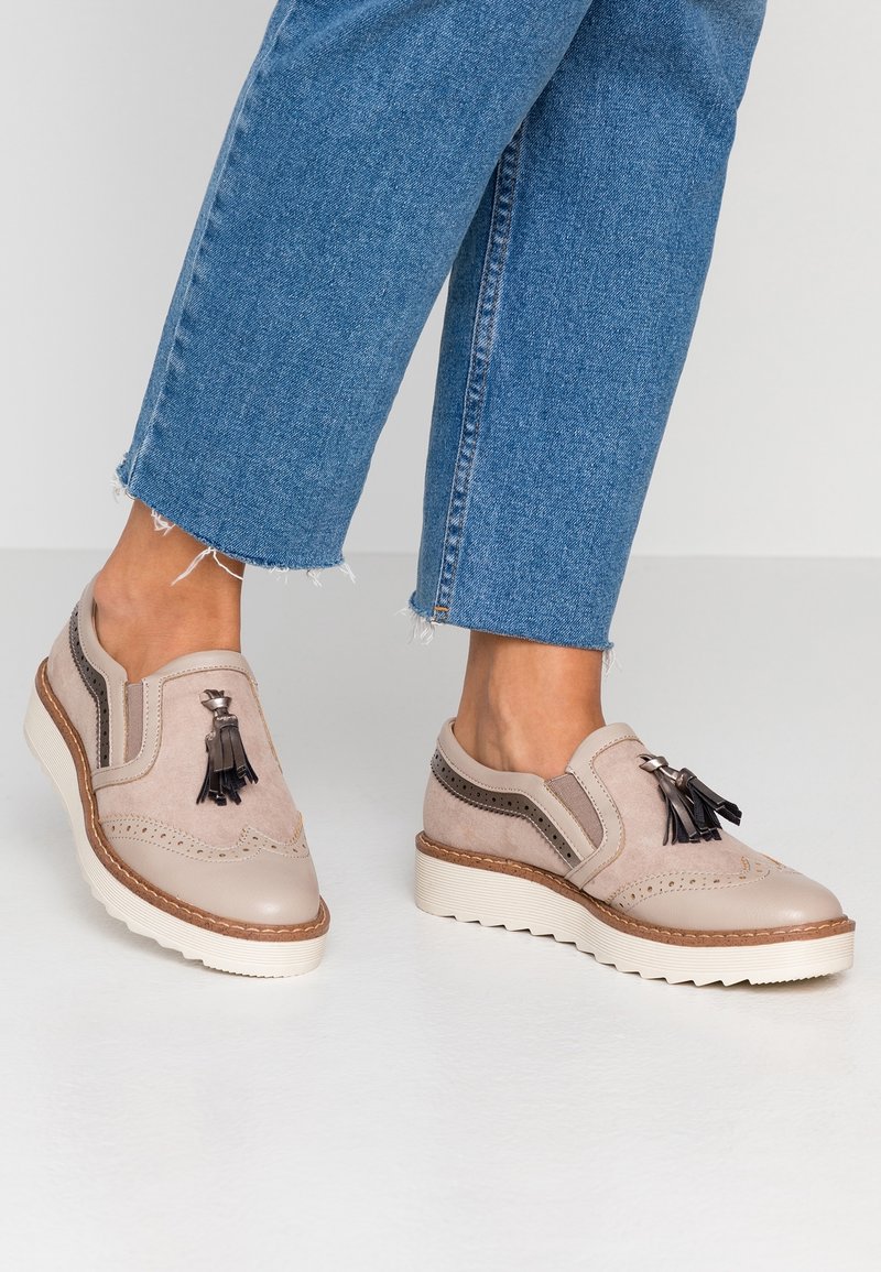 Chaussures en cuir beige et suède avec des pampilles décoratives et des accents perforés. Semelle blanche épaisse ; portées avec un jean en denim effiloché.