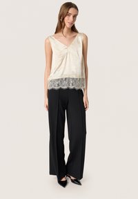 Top senza maniche di colore crema chiaro con scollatura a V e tessuto texture, caratterizzato da un orlo in pizzo. Abbinato a pantaloni a vita alta neri a gamba larga e scarpe con punta.