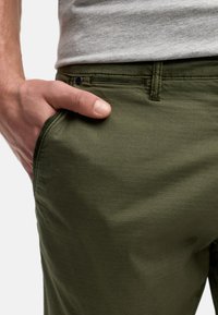 Zelene hlače od glatke tkanine, slim fit kroja, s bočnim džepom i suptilnom teksturom. Pojas ima mali plavi naglasak.