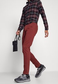 En person som bär en svart och röd rutig skjorta, röda byxor och svarta sneakers, håller en liten svart väska och går mot en enkel bakgrund.