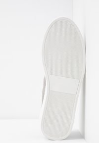Semelle blanche de basket avec des rainures horizontales, placée verticalement contre un mur blanc d’angle sur une surface blanche.