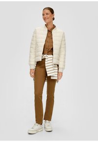 Veste matelassée crème, chemise à boutons marron, pantalon marron et baskets blanches. Présente une écharpe rayée nouée à la taille. Texture lisse, design ajusté.