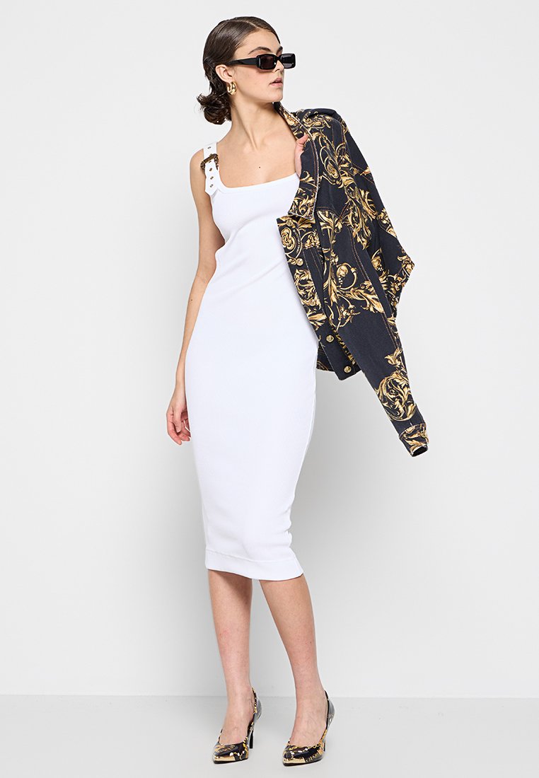 Vestido blanco ajustado con escote cuadrado y botones decorativos en los hombros, combinado con una chaqueta negra y dorada con estampado y tacones estampados.