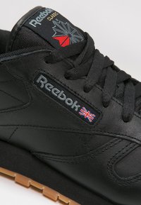 Svart lær sneakers med gummisåle, med Reebok-logo og Union Jack-emblem på tungen, teksturerte snørebånd og diskrete sømdetaljer.