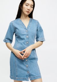 Νεαρή γυναίκα με μακριά σκούρα μαλλιά, ντυμένη με εφαρμοστό φόρεμα από τζιν με κοντά μανίκια και κουμπιά στο μπροστινό μέρος, στέκεται με δεμένα τα χέρια.