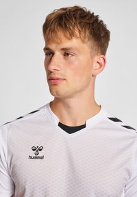 Hvid sportsjersey med et struktureret mønster, sorte detaljer på skuldrene og en V-halsudskæring. Hummel-logoet præsenteret i sort på fronten.