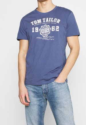 Granatowa koszulka z krótkim rękawem i okrągłym dekoltem, z białym nadrukiem tekstu i logo, w połączeniu z jasnoniebieskimi dżinsami.