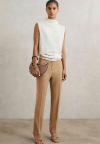 Top senza maniche color beige chiaro con un colletto alto, abbinato a pantaloni beige su misura e sandali con cinturini beige, mentre tiene una borsa marrone.
