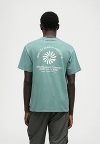 T-shirt verde din bumbac, cu mâneci scurte, având un design grafic alb pe spate, care include text și un motiv floral.