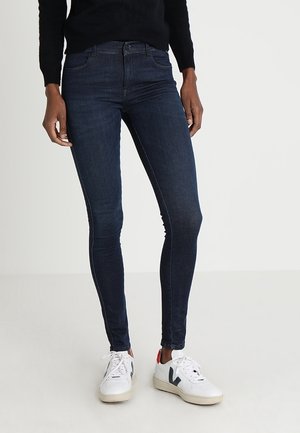 Person wearing dark blue skinny jeans, white sneakers with black and red accents, and a black long-sleeve top against a plain light background.