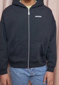 Personne portant un sweat à capuche noir zippé avec le texte "WRSTBHVR" sur la poitrine et un jean bleu clair, debout devant un fond rose.