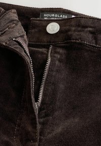 Brązowe spodnie z korduroyu z metalowym guzikiem i suwakiem, charakteryzujące się fakturowaną powierzchnią i luźnym krojem. Metka głosi "HOURGLASS RELAXED BOOT."