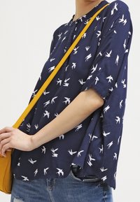 Blouse bleu marine avec motifs d'oiseaux blancs, manches courtes, col rond, coupe décontractée, associée à un sac bandoulière jaune.