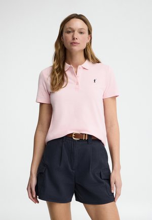 Kobieta w jasnoróżowej koszulce polo z krótkim rękawem, wpuszczonej do czarnych krótkich spodenek typu cargo z wysokim stanem i brązowym paskiem.