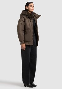 Brun polstret jakke med høj krave, lynlås foran og sidelommer, båret over en sort top og parret med mørke brede bukser og sorte sko.