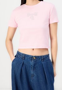 Camiseta corta rosa de mangas cortas con un diseño de lazo de strass en el pecho, combinada con pantalones de mezclilla de cintura alta.