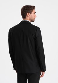 Musta räätälöity pikkutakki, jossa on jämäkkä siluetti, lovitetut kaulukset ja sileä pinta; sisältää napilla varustetut hihansuut ja viilto takana.