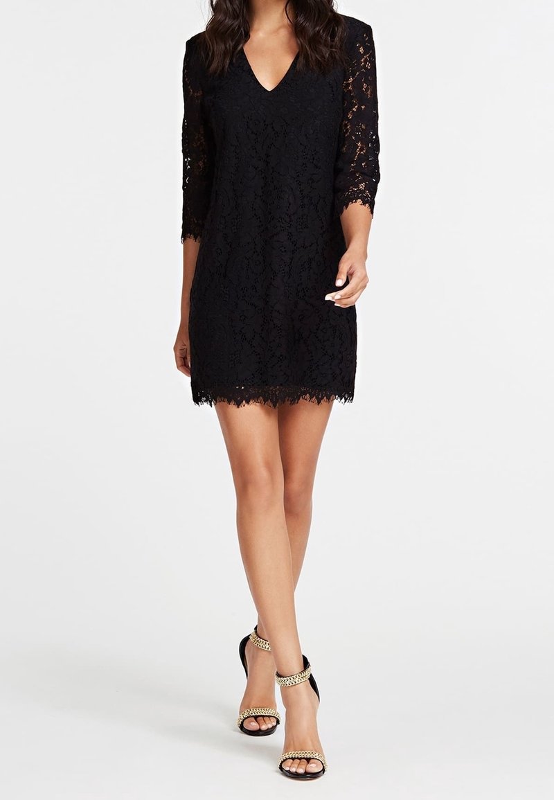 Femme portant une robe noire en dentelle à col en V avec des manches trois-quarts et des talons hauts noirs avec des brides chaîne dorées, marchant vers l’avant.