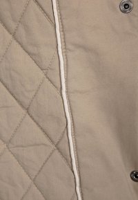 Beige vävda och släta tygpaneler sammanfogade med en vit söm, med en metalltryckknapp på den släta tygsidan.