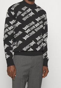 Svart crewneck-tröja med vit "Just Cavalli" text tryckt diagonalt över hela, bärs med grå byxor.