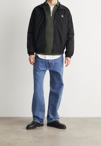 Blouson aviateur noir avec un pull vert en maille en dessous, une chemise blanche apparaissant, un jean léger bleu clair ample et des chaussures noires.