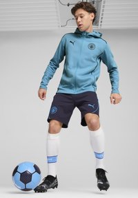 Lyseblå lynlåsejakke med hætte og sorte detaljer, marineblå shorts med blå dekoration, hvide sokker, sorte fodboldstøvler og en blå bold.