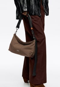 Bolso de hombro marrón con textura suave, con diseño de solapa, correa negra ajustable y marca sutil en la esquina inferior.