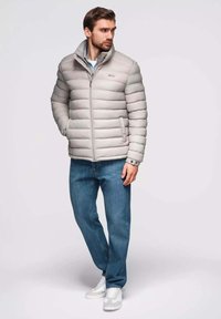 Grau gefütterte Jacke mit horizontalem Steppmuster, Frontreißverschluss und Seitentaschen, kombiniert mit blauen Jeans und weißen Sneakers.