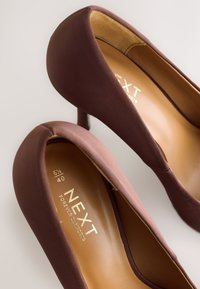 Brune højhælede sko med en glat, mat stofoverflade og rundet tå. Indvendigt har de en beige indersål med mærkelogo.