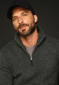 Pull gris zippé avec un tricot texturé, présentant un col haut. Accompagné d'un T-shirt gris et d'une casquette noire. Fond neutre.