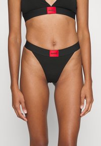 Čierny bikinový set obsahujúci tangá s nízkym pásom a braletku so širokými ramienkami. Obe kusy majú červené logo "HUGO" na páse a na poprsí.