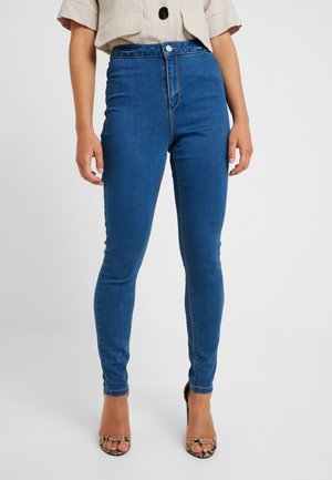 Vrouw draagt hoge taille blauwe skinny jeans gecombineerd met beige cropped top en open teen hakken met slangenprint, staand op een witte achtergrond.