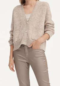 Femme portant un cardigan beige boutonné avec deux poches frontales sur un haut beige, associé à un pantalon taupe brillant, une main dans la poche.