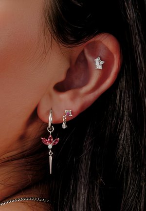 Ear with three silver earrings: a dangling one with pink gems shaped like petals, a small stud with a purple gem, and a star-shaped stud.