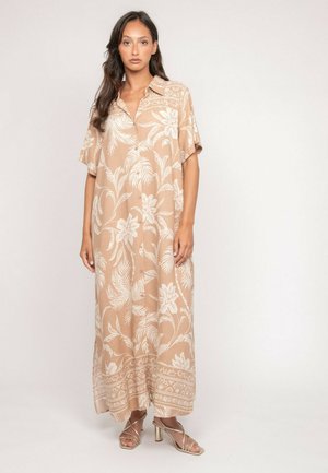 Femme debout, portant une robe maxi beige ample à imprimé floral blanc, manches courtes, col et sandales à talons hauts métalliques. Fond neutre.