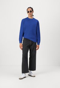 Blue knitted sweater with textured pattern, paired with dark wide-leg jeans and white sneakers. Black round sunglasses complete the look.
